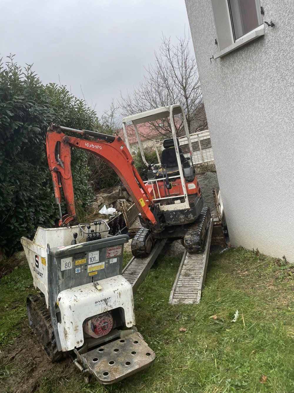 Mini-pelle et plaque vibrante acheminées sur rampes dans un accès étroit le long d'une maison
